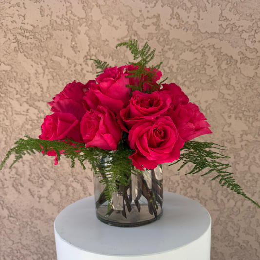 Bouquet of pink roses in a clear vase on a white pedestal against a beige textured wall.
