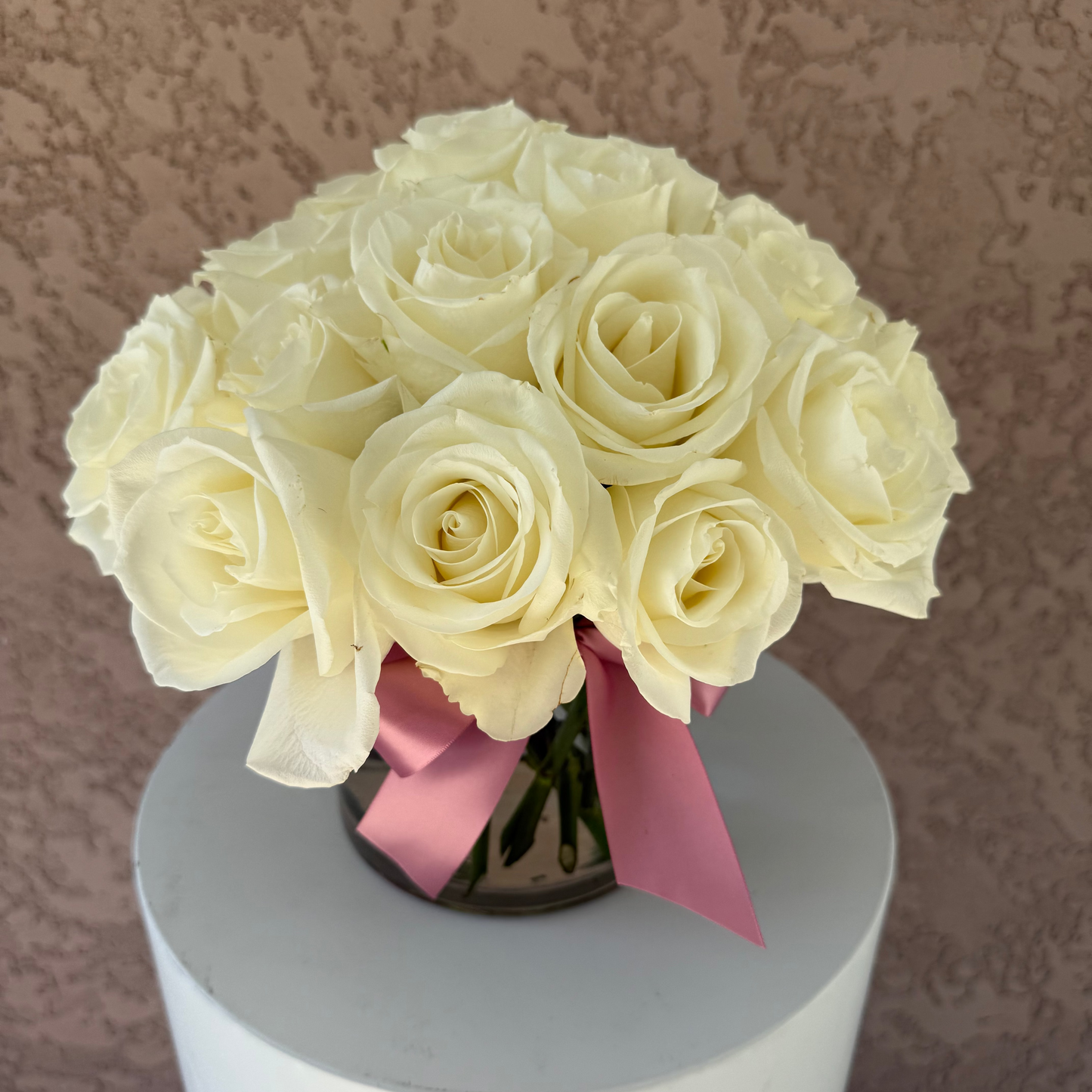 Bouquet of white roses with a pink ribbon in a clear vase on a white surface.