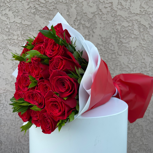 Bouquet of red roses wrapped in white paper with a red ribbon against a beige wall.