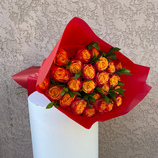 Bouquet of orange roses wrapped in red paper and placed in a white vase against a textured wall.