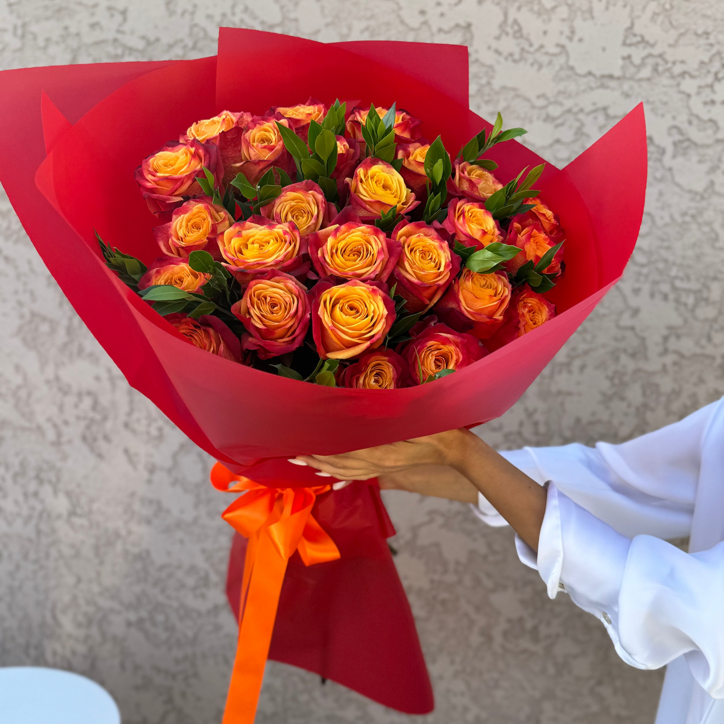 Bouquet of red and orange roses in red paper with an orange ribbon, held by a person in a white sleeve.
