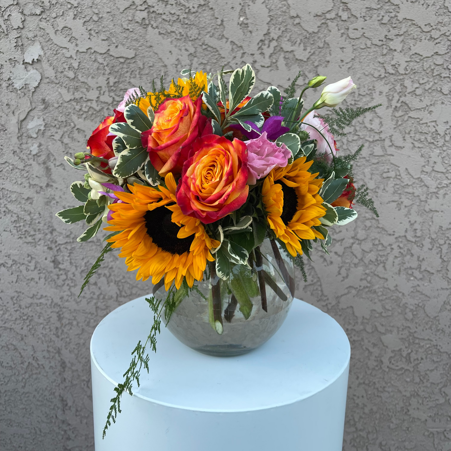 Colorful flower arrangement with sunflowers and roses on a white pedestal against a gray textured wall.