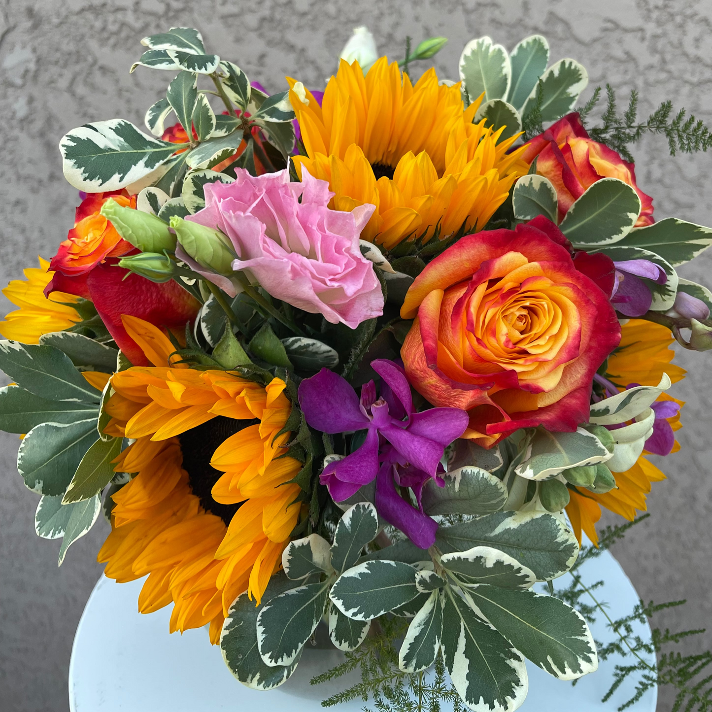 Colorful flower bouquet with sunflowers, roses, and other flowers in a vase against a gray wall.