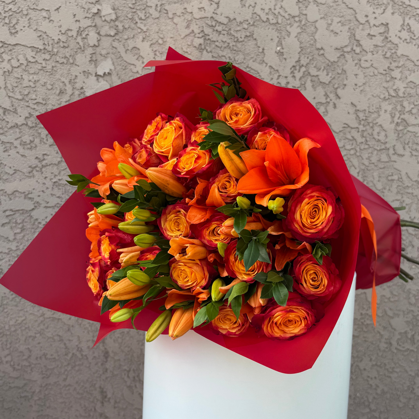 Bouquet of orange flowers wrapped in red paper on a white pedestal against a textured gray background