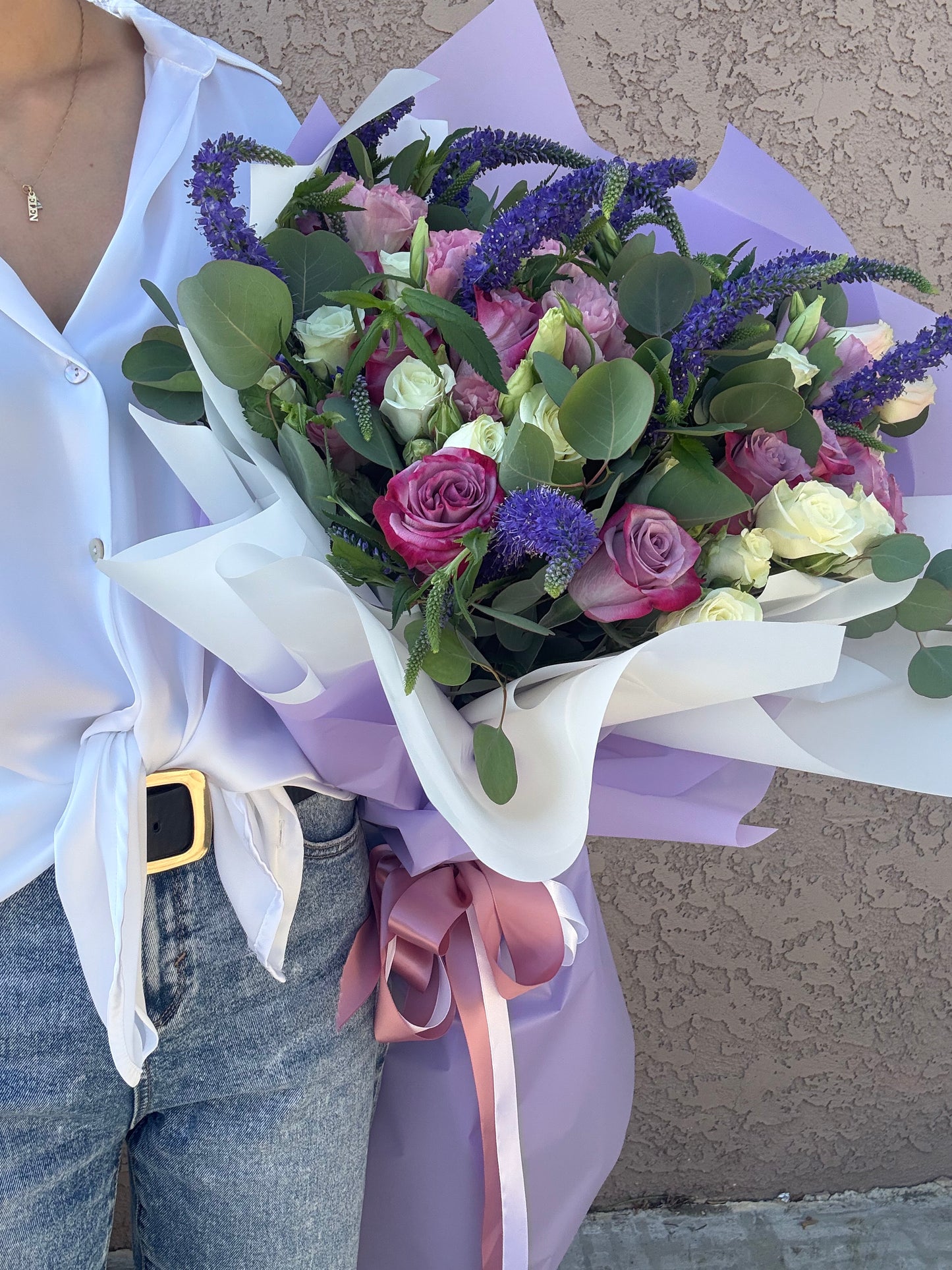 Bouquet of flowers wrapped in white paper with a pink ribbon, held by a person wearing a white shirt and blue jeans.