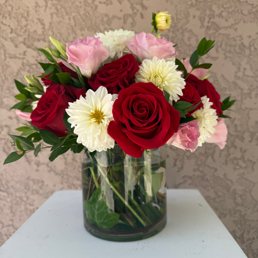 Bouquet of red, pink, and white flowers in a clear vase on a light surface with a textured beige background.