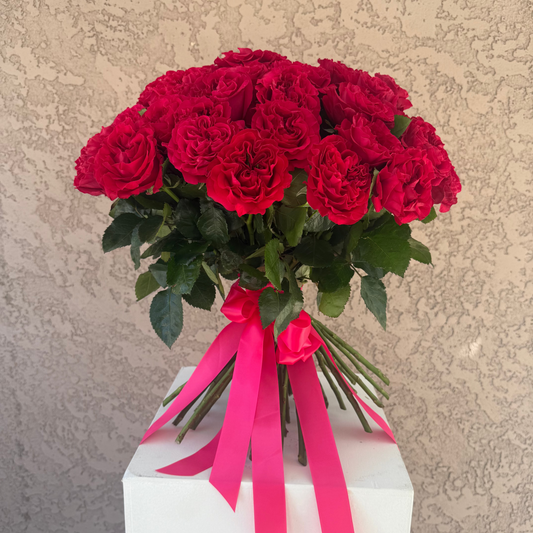 Bouquet of red roses with pink ribbons on a white pedestal against a beige wall.