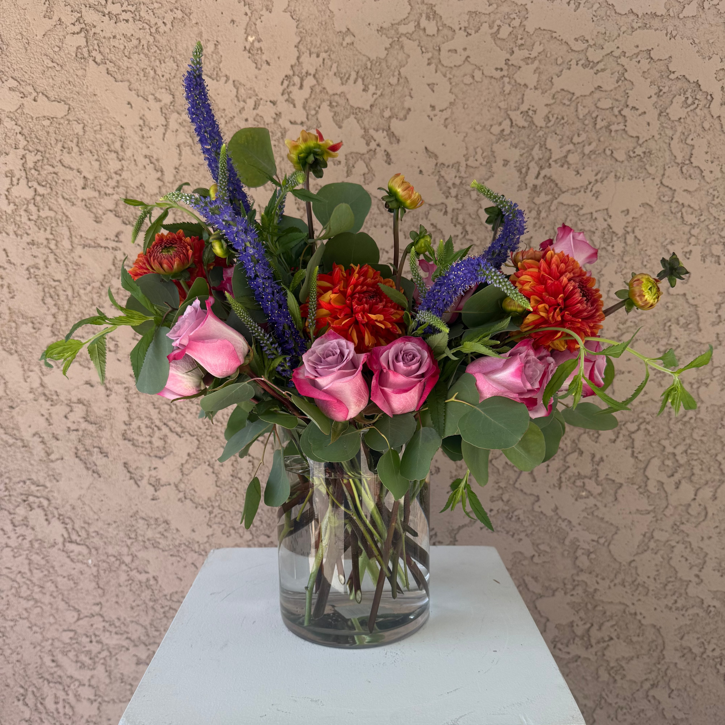 Bouquet of colorful flowers in a clear vase on a white pedestal against a beige wall.