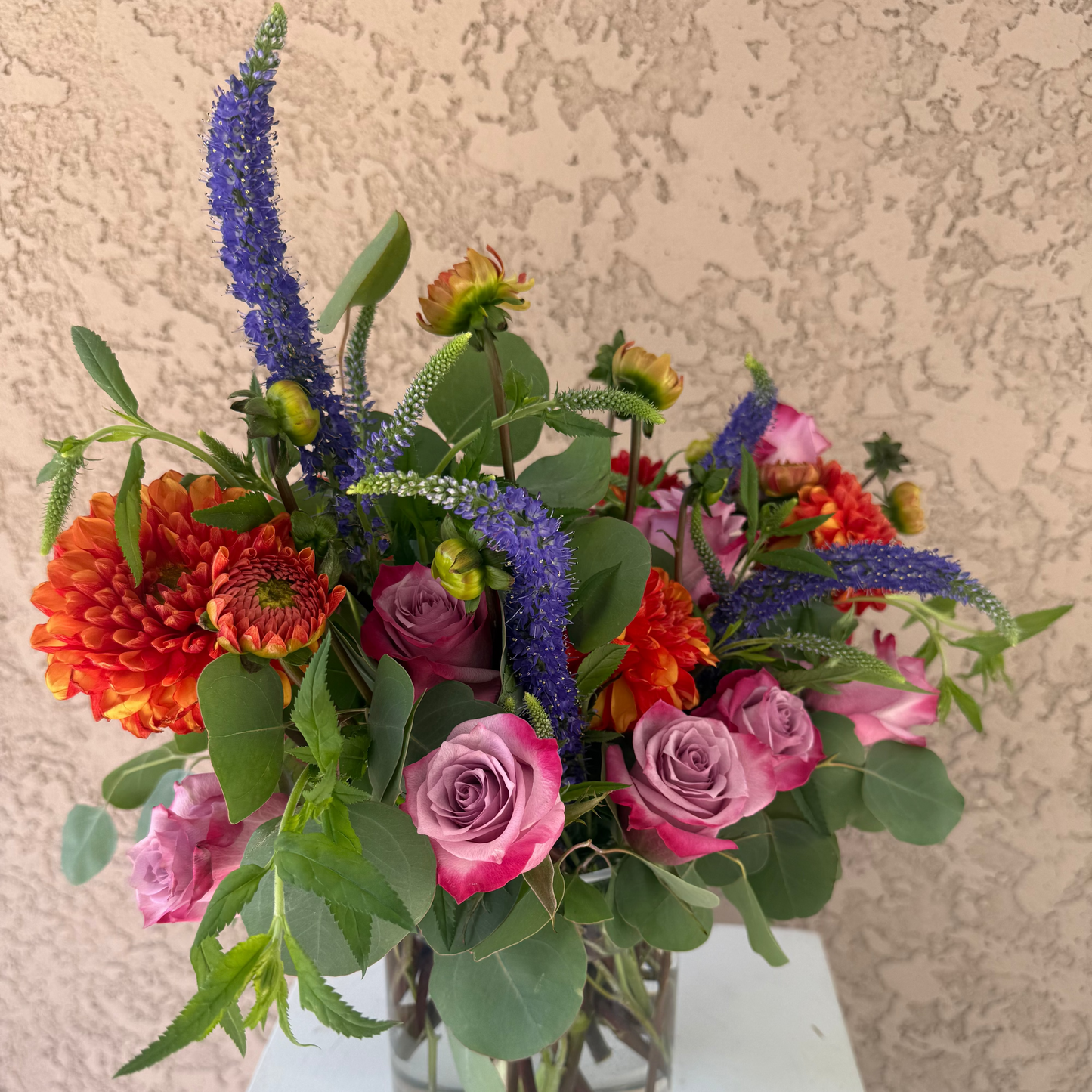 Colorful flower arrangement in a glass vase on a white surface with a textured beige wall background
