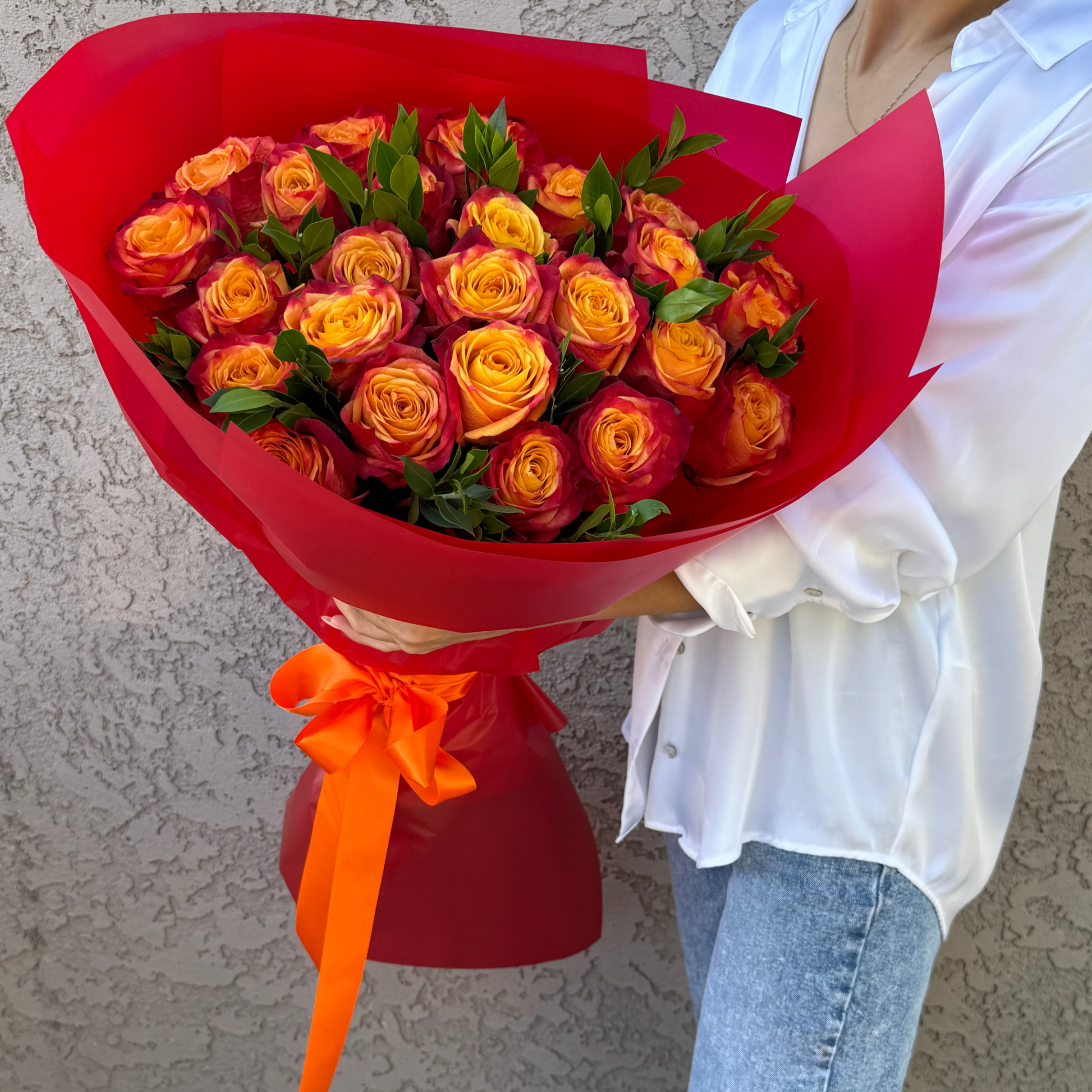 Bouquet of orange roses in a red wrap held by a person wearing a white shirt and blue jeans.