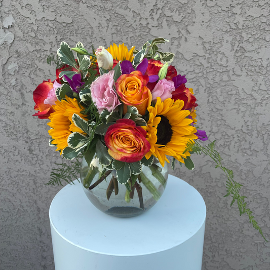 Colorful flower arrangement in a clear vase on a white pedestal against a textured gray wall.