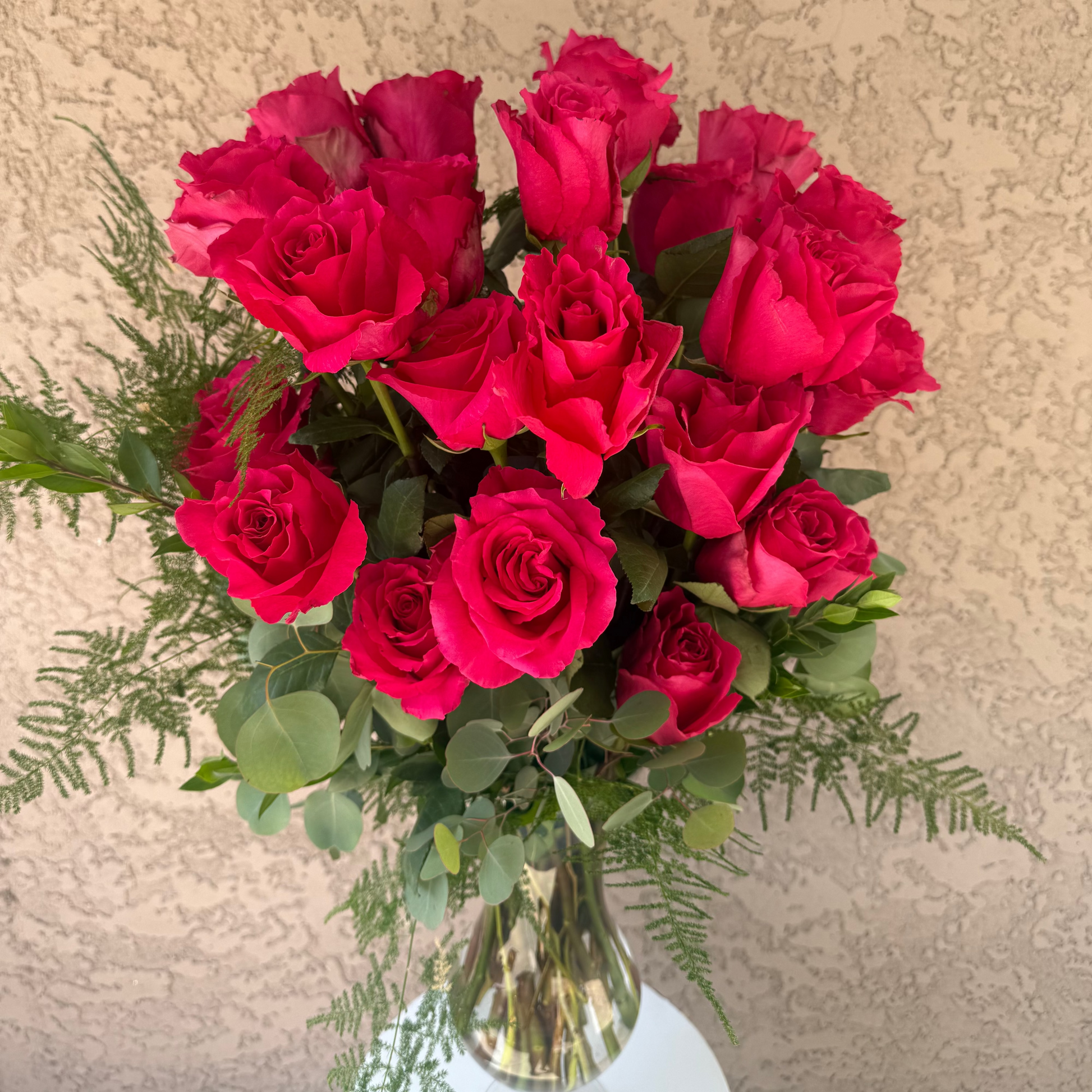 Bouquet of red roses with greenery in a clear vase against a beige textured wall.