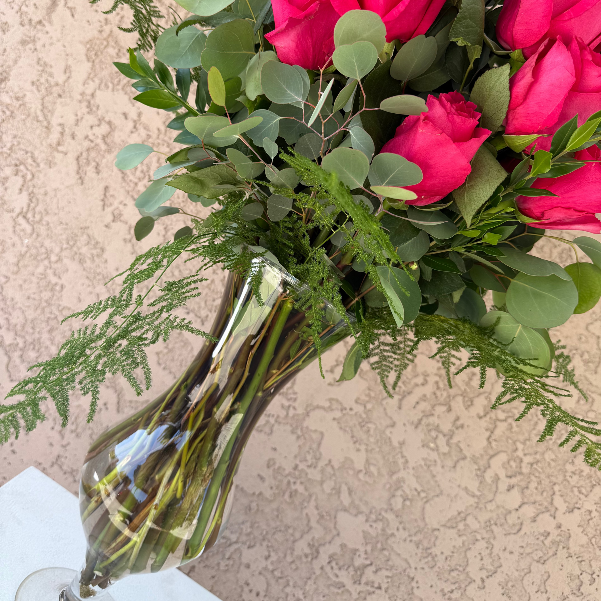 Bouquet of pink roses with greenery in a clear vase against a beige wall.
