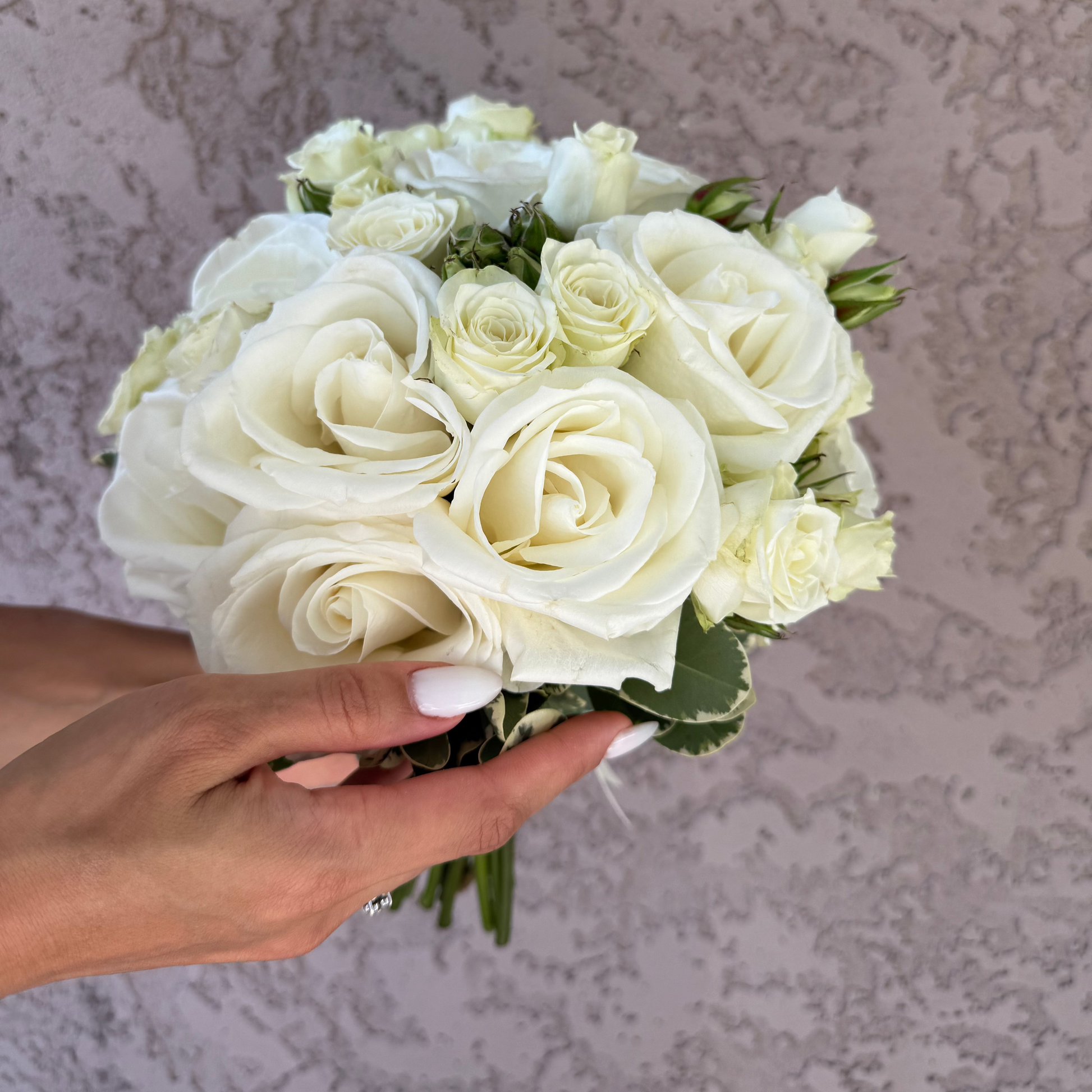 Bouquet of white roses held by a hand against a textured beige wall.
