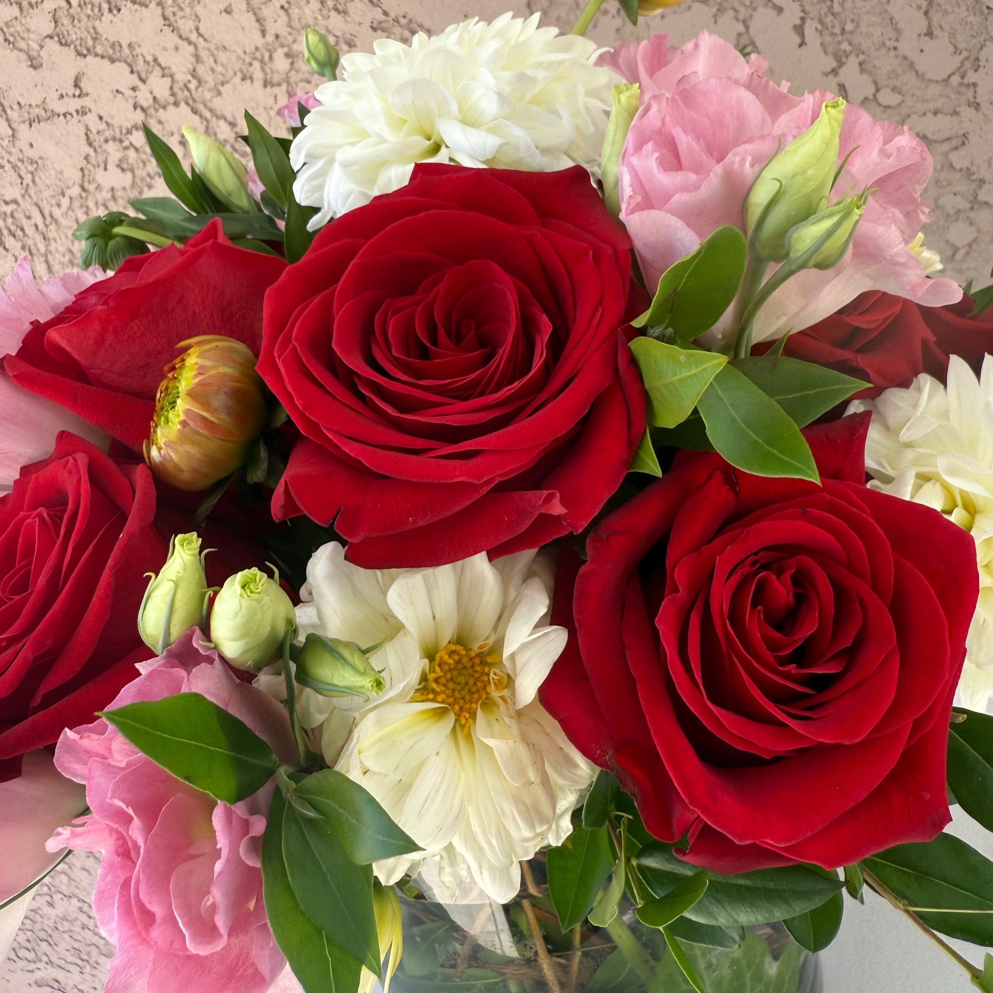Bouquet of red roses, white flowers, and pink roses in a clear vase against a neutral background.