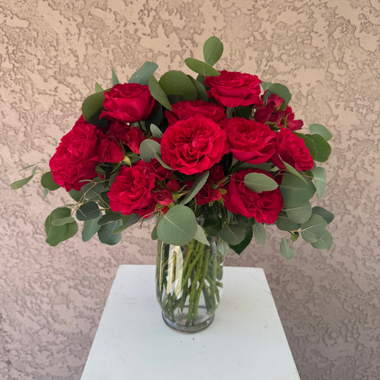 Bouquet of red roses in a clear vase on a white pedestal against a beige wall.