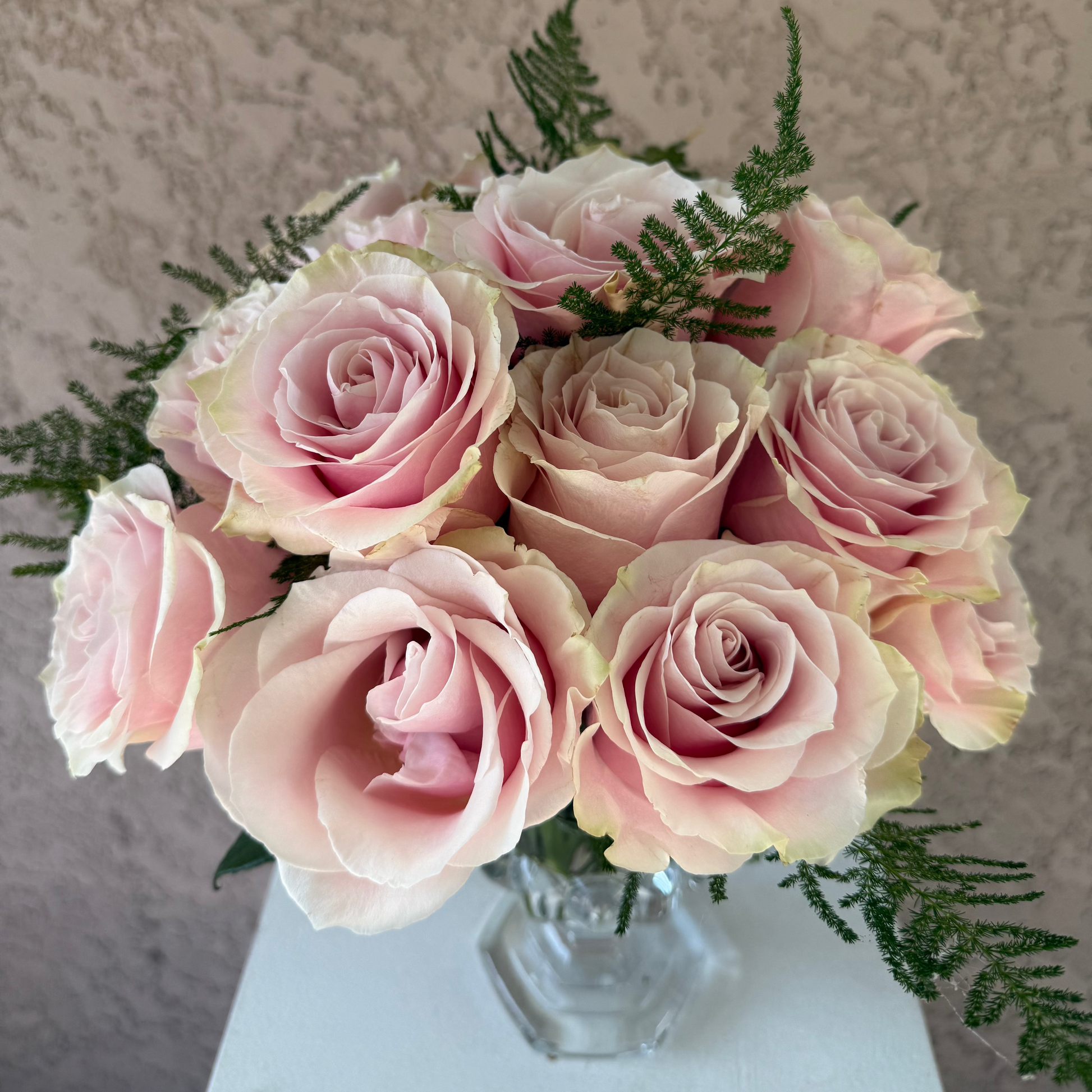 Bouquet of pink roses in a clear vase against a textured beige wall.