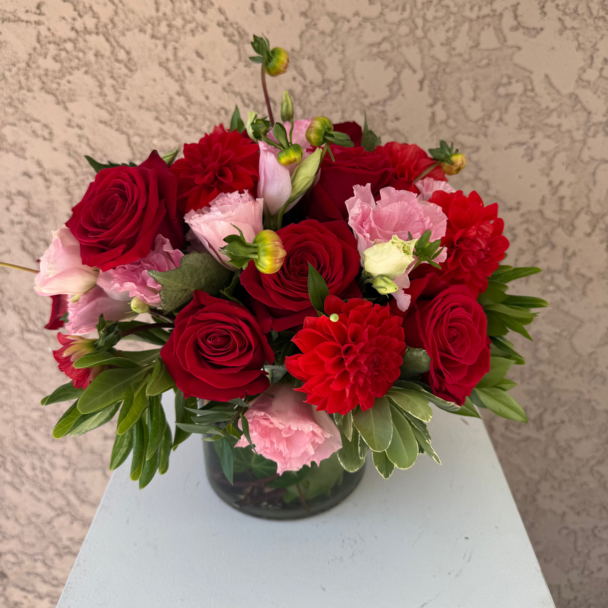 Bouquet of red and pink flowers in a clear vase on a light surface with a textured beige wall background.