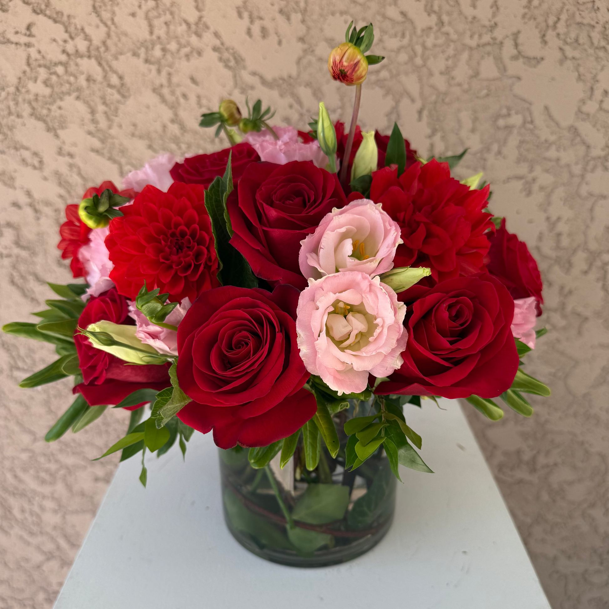 Bouquet of red and pink flowers in a clear vase on a white surface with a beige textured background