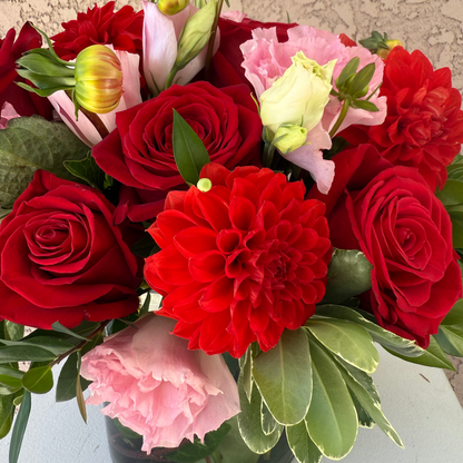 Bouquet of red roses and dahlias with pink flowers in a clear vase against a textured wall.