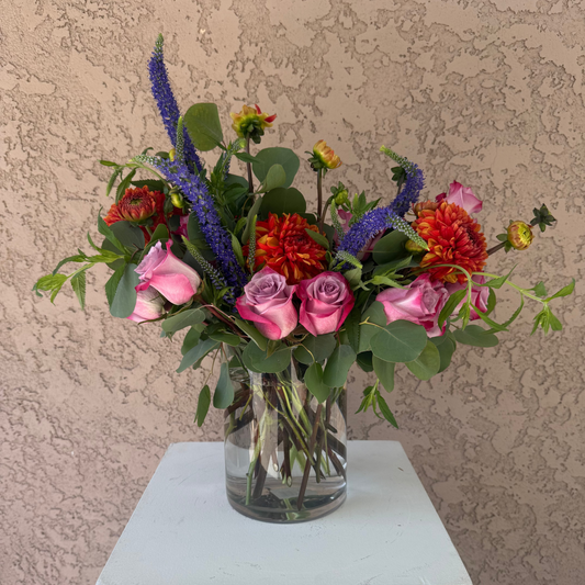 Bouquet of colorful flowers in a clear vase on a white pedestal against a beige wall.